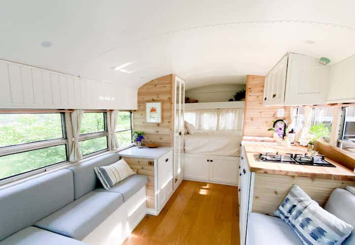 The interior of the bus is light and airy, with soft gray Sunbrella upholstered bench seating, natural wood finishes, a cozy kitchen, and a nook for a full-size bed.