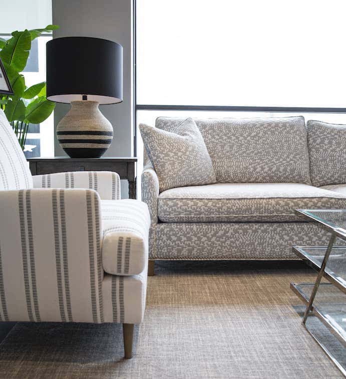 Seating area with sofa upholstered in a gray and white patterned Sunbrella performance fabric, end chair upholstered in a striped Sunbrella fabric, glass coffee table and accessories found at the CR Laine showroom.